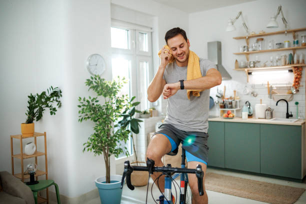 Man checks smartwatch while riding stationary bike at home, illustrating wearable tech in fitness routines.