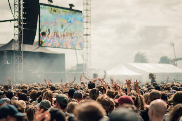 A crowd at an outdoor festival joyfully raises their hands, with a large screen displaying colorful visuals.